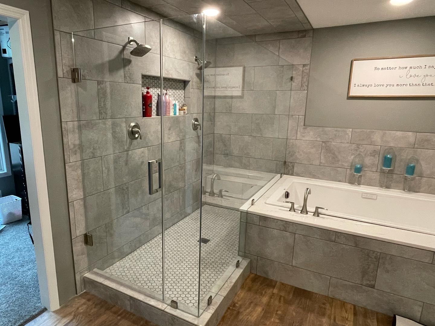 Frameless corner shower enclosure with a swing door, brushed hardware and grey stone-look tile next to a soaking tub